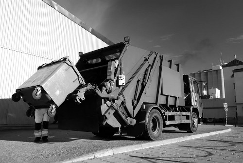A black and white photograph depicting a large commercial rubbish collection vehicle parked on a paved urban street near a warehouse or industrial building. The vehicle is equipped with hydraulic arms that are in the process of lifting a large wheeled waste bin, which has a textured, slightly rough surface and is fitted with sturdy wheels and a metal handle. The bin appears to be made of plastic and is filled with waste materials, with some debris visible at the opening. To the left of the vehicle, a worker dressed in high-visibility clothing is standing beside the bin, guiding it into the truck's loading compartment. In the background, there is a tall corrugated metal wall, typical of industrial sites, and a partly cloudy sky overhead. The scene captures the active process of rubbish removal on a commercial or industrial premises, aligning with the services offered by Commercial Waste Removal Sydenham in South London, near postcode SE26, in a professional and factual manner.