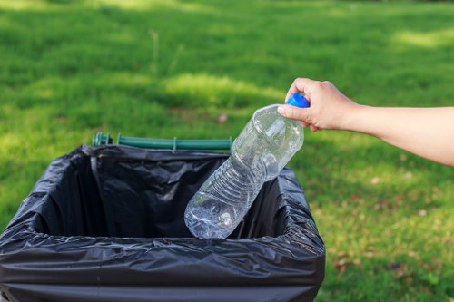 A person's hand is seen holding a clear plastic water bottle with a blue cap, poised above an open black plastic rubbish bin on a grassy outdoor area. The bin is positioned on a paved surface, and the background features a well-maintained green lawn with blurred foliage. The water bottle appears partially crushed, with visible ridges and transparency showing the liquid level inside. The scene is lit by natural daylight, emphasizing the textures of the plastic bottle and the smooth, shiny surface of the bin. This image relates to waste disposal and rubbish removal services offered by Commercial Waste Removal Sydenham, capturing the act of discarding household waste in an outdoor setting, typical of the type of rubbish collected in the Sydenham postcode area.