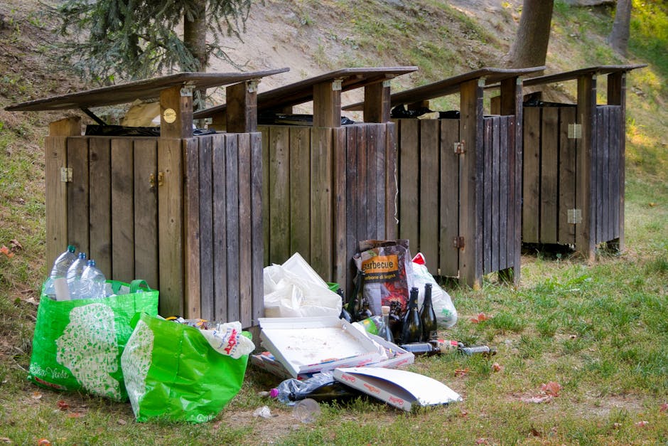 An outdoor scene shows four wooden public trash bins placed on a grassy area, each with a slanted roof, weathered and stained with dark brown and grey finishes. In the foreground, discarded rubbish is