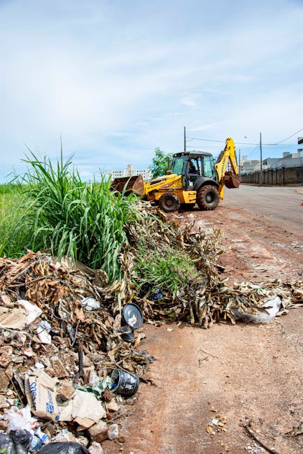 A waste collection vehicle with an open rear compactor area, positioned on a narrow urban street lined with older multi-story buildings. The truck is white and appears to be in the process of emptying
